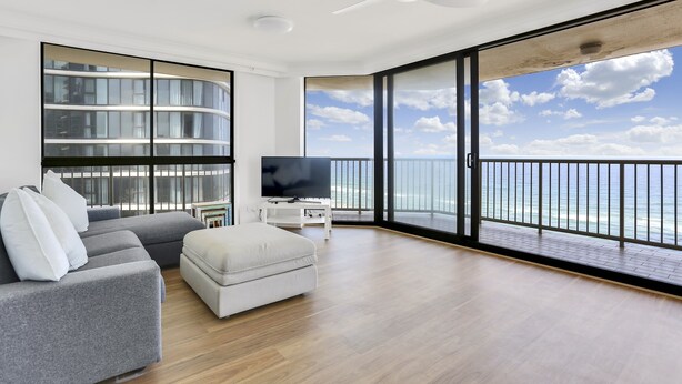 A modern living room with a grey sectional sofa, a white ottoman, and a flat-screen TV on a white stand. A large window offers a view of the ocean and a balcony with a railing.
