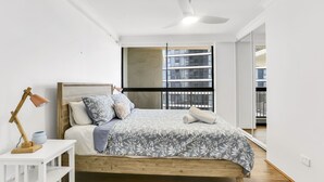 A modern bedroom with a wooden bed, a white nightstand, a floor lamp, and a large window with a view of buildings.