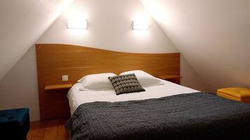 Una habitación pequeña y triangular con cabecera de madera, cama con almohadas blancas y estampadas, y colcha oscura. Hay dos lámparas fijadas a la pared y una mesita de noche de madera con un cojín amarillo.