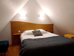 Una habitación pequeña y triangular con cabecera de madera, cama con almohadas blancas y estampadas, y colcha oscura. Hay dos lámparas fijadas a la pared y una mesita de noche de madera con un cojín amarillo.