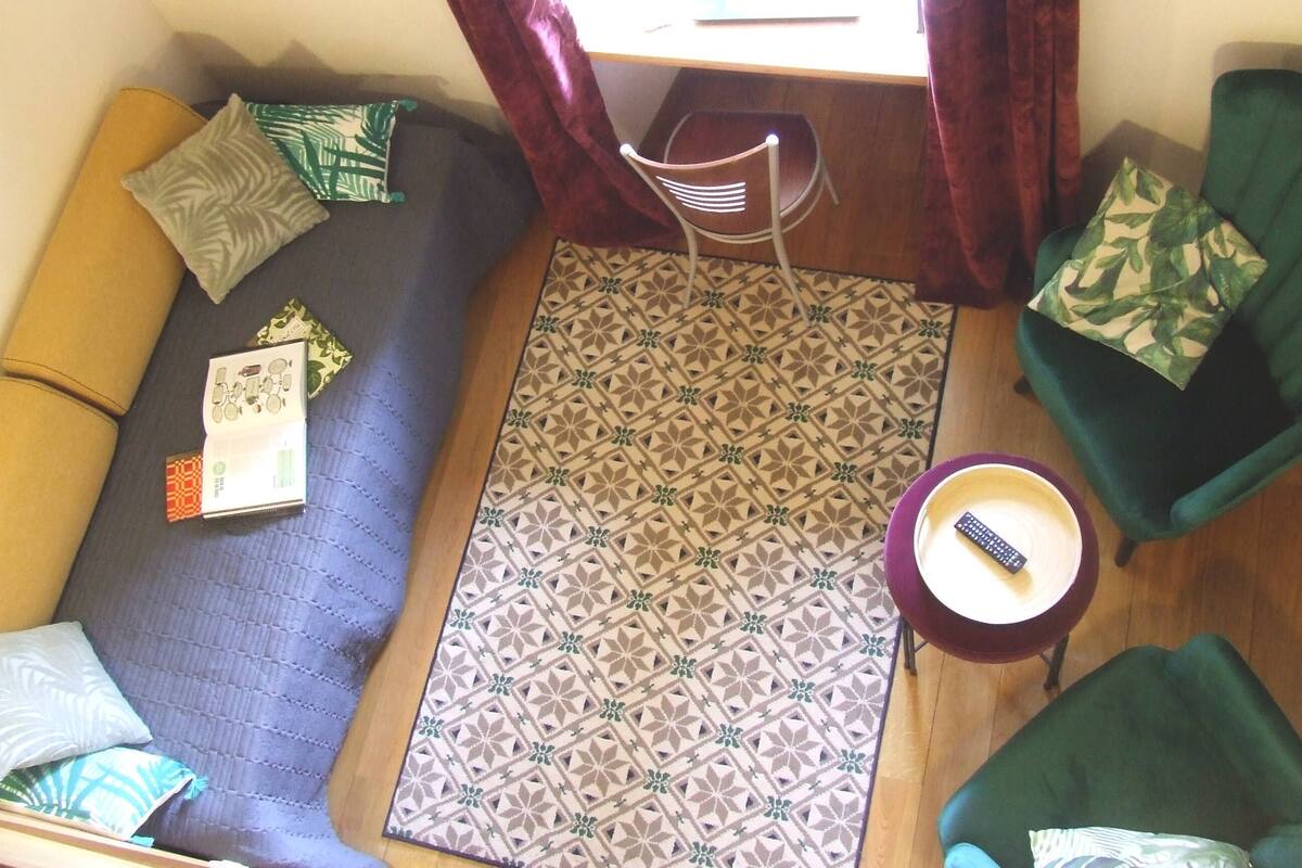 Una habitación con una alfombra estampada, un sofá, una silla, una mesita con un control remoto y una computadora portátil sobre un escritorio.