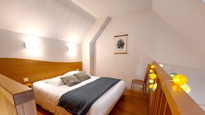 Une chambre à coucher avec une tête de lit en bois, un lit recouvert de draps blancs et d’une couverture grise, une petite table de chevet, une chaise et un luminaire fixé au mur.