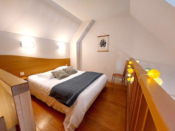 Un dormitorio con cabecera de madera, cama con ropa blanca y una manta gris, una mesita de noche pequeña, una silla y un aplique de pared.