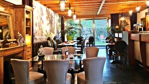 Un restaurant avec des poutres en bois au plafond, des luminaires suspendus et une vue sur la verdure environnante. Les tables sont disposées avec des chaises, et des objets décoratifs sont placés sur des étagères.