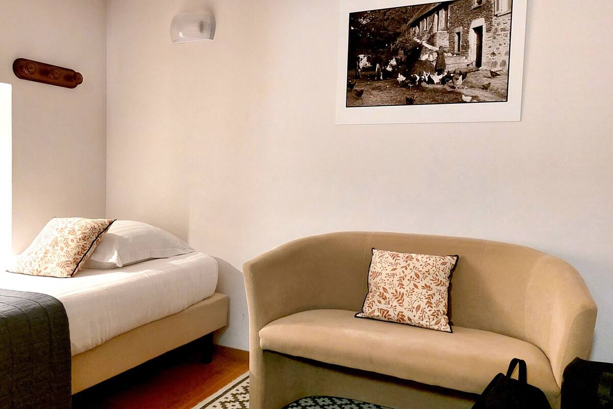 Un dormitorio con cama, silla, almohada, luz empotrada en la pared y un cuadro enmarcado.