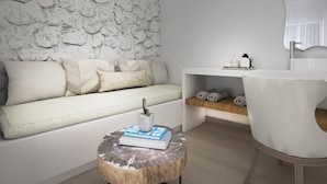 Una sala moderna con pared de textura de piedra, un sofá blanco con cojines estampados, una mesita de madera con un libro, una silla blanca y un mueble consola blanco con toallas y una pequeña planta.