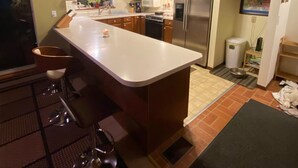 Una cucina con mobili in legno, un frigorifero in acciaio inossidabile, una grande isola con sgabelli e una parete di fondo rivestita di piastrelle.