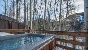 Un patio in legno con una vasca idromassaggio circondata dalla neve, con vista su una zona boschiva e montagne sullo sfondo.