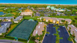 An aerial view of a coastal resort with a tennis court, parking areas, and beachfront properties.