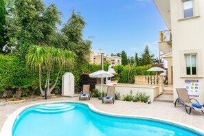 Piscina com espreguiçadeiras e guarda-sóis, rodeada por vegetação e um edifício com área de lazer à beira da piscina.