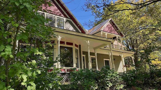 A two-story house with a porch, surrounded by trees and shrubs. - Your Beautiful Victorian Getaway (Bethel)