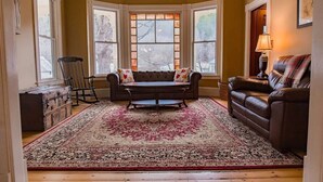 A cozy living room with a brown leather sofa, a wooden coffee table, and a patterned rug. There are three windows with white frames and a lamp on a side table. - Your Beautiful Victorian Getaway (Bethel)