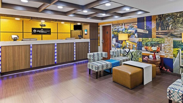 A hotel reception area with a wooden reception desk, seating area with patterned chairs and ottomans, and a mural on the wall depicting various scenes. - Clarion Pointe Savannah Gateway I-95 (Savannah)