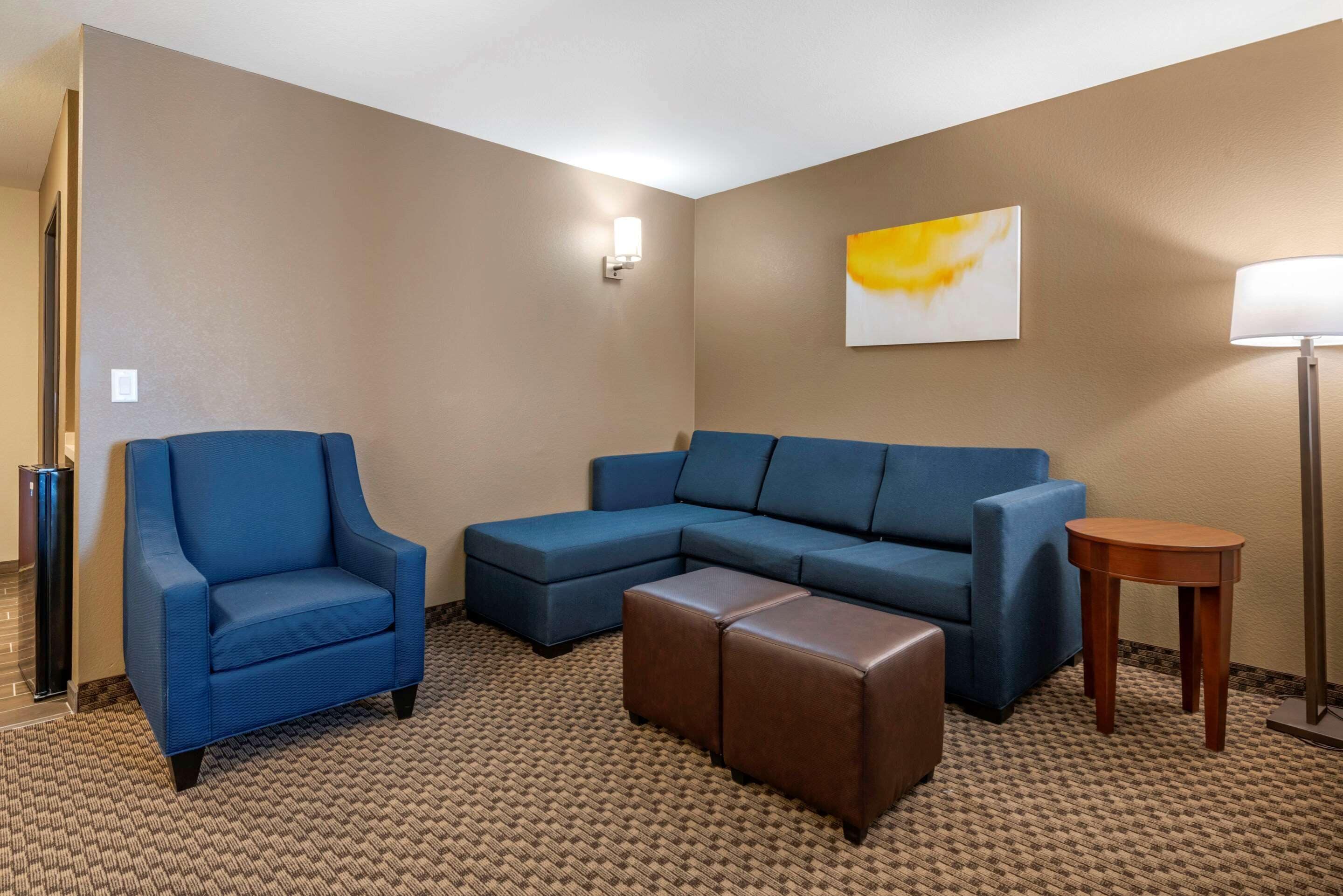 Quarto de hotel com uma poltrona azul, um sofá de canto e uma pequena mesa lateral de madeira.