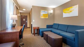 A hotel room with a wooden desk, a blue sofa, an ottoman, a small round table, and a lamp.