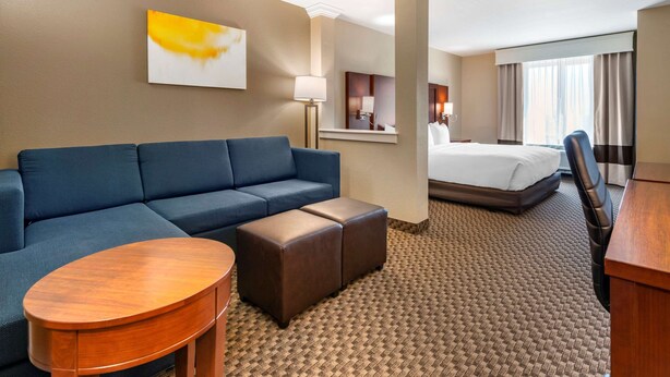 A hotel room with a blue sofa, a wooden coffee table, a bed with white linens, and a large window with curtains. - Comfort Suites Broomfield-Boulder/Interlocken (Broomfield)