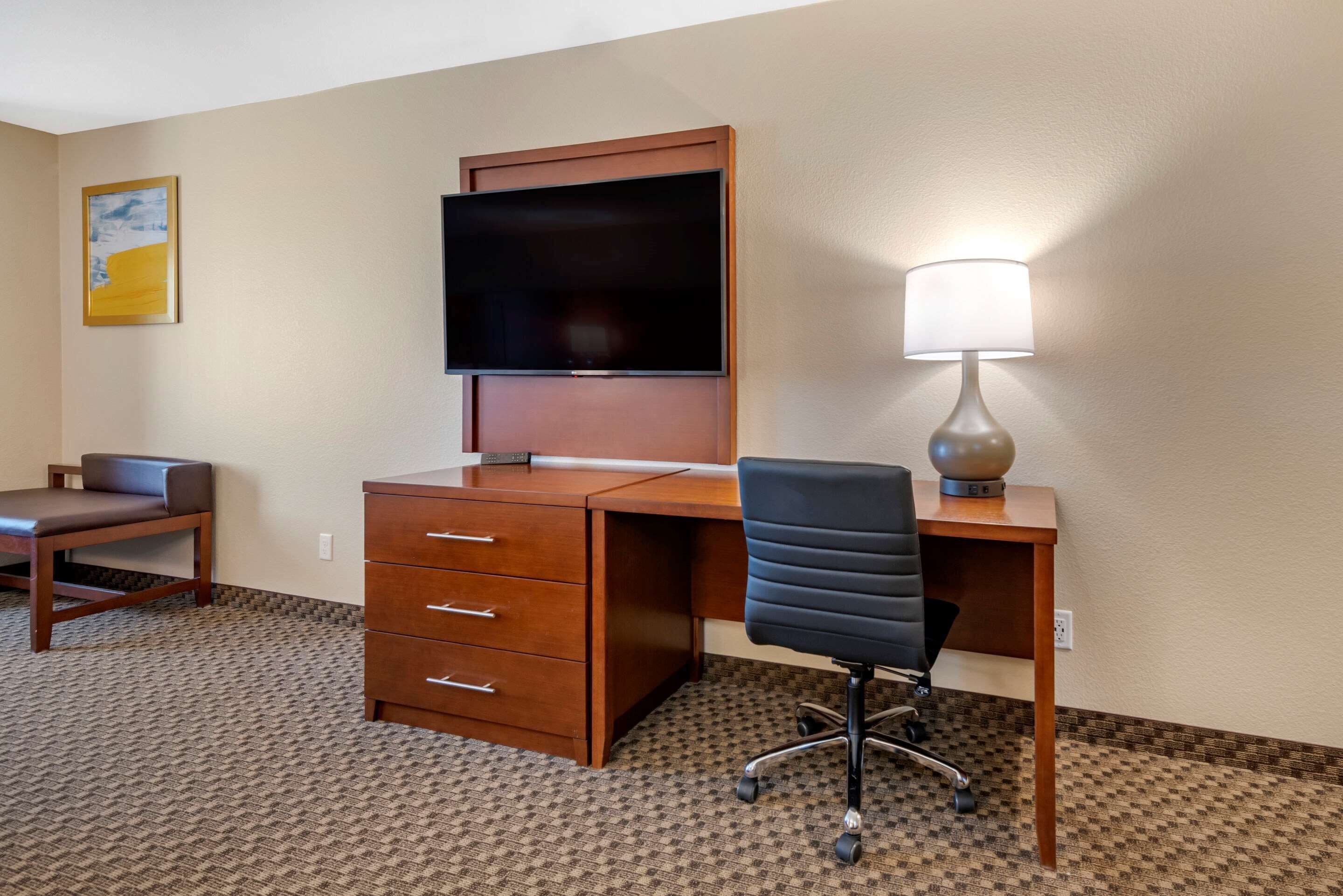 A hotel room with a wooden desk, a black office chair, a flat-screen TV mounted on the wall, a lamp on the desk, and a small sofa.