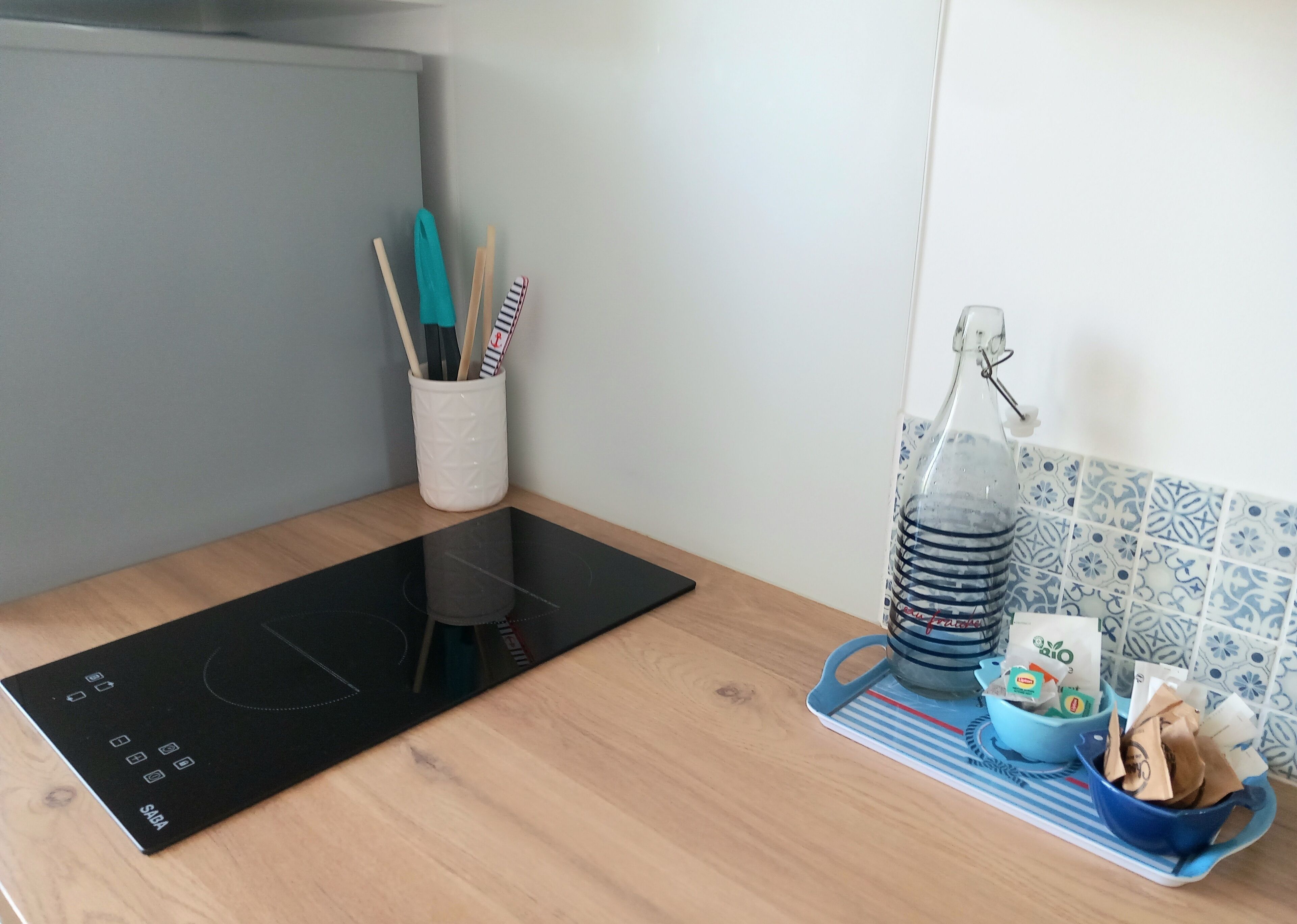Una cocina con un fogón de inducción negro, un portalapilas blanco con utensilios y un cubo de basura azul con bolsa para reciclaje.