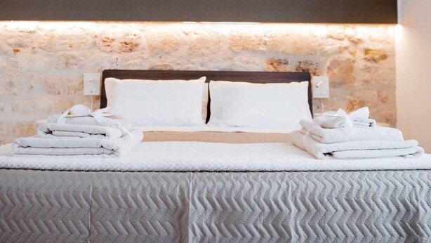 Un lit bien fait, avec du linge de lit blanc et des oreillers, une couvre-lit blanche à motifs et un mur en pierre derrière le lit.