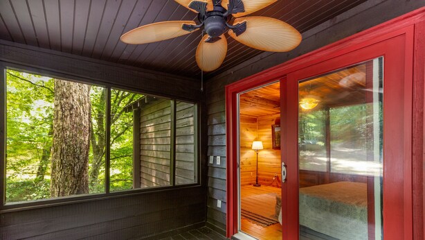 Une terrasse en bois avec un ventilateur de plafond, une porte rouge donnant sur une pièce éclairée par une lampe, et une vue sur les arbres à travers de grandes fenêtres.