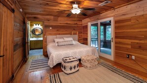 Une chambre située dans une cabane en bois, avec un lit, un ventilateur de plafond, une grande fenêtre et une porte donnant sur un balcon.