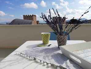 A table with a white tablecloth, a polka-dotted cup, a spoon, and a vase with lavender on a rooftop terrace with a view of a castle and mountains.