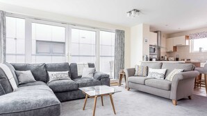 A modern living room with grey sofas, a glass coffee table, and a kitchen in the background. - Steep Holm - With Spacious Balcony (Poole)