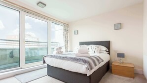 A bedroom with a bed, bedside table, lamp, and a view through a window. - Steep Holm - With Spacious Balcony (Poole)