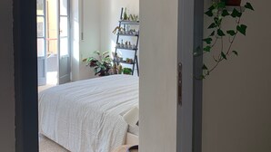 Un dormitorio con una cama, un taburete de madera, una estantería con plantas y vistas al exterior.