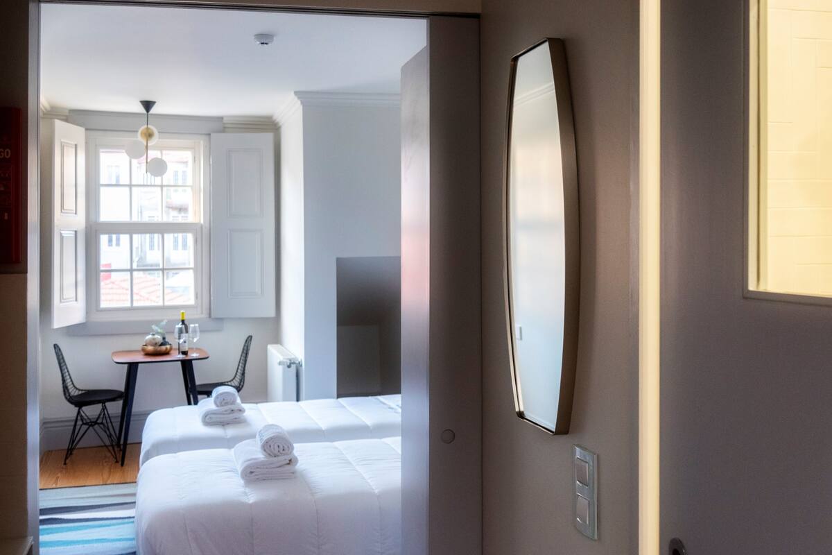 A hotel room with two beds, a small table with a bottle and glass, a chair, and a large mirror on the wall.