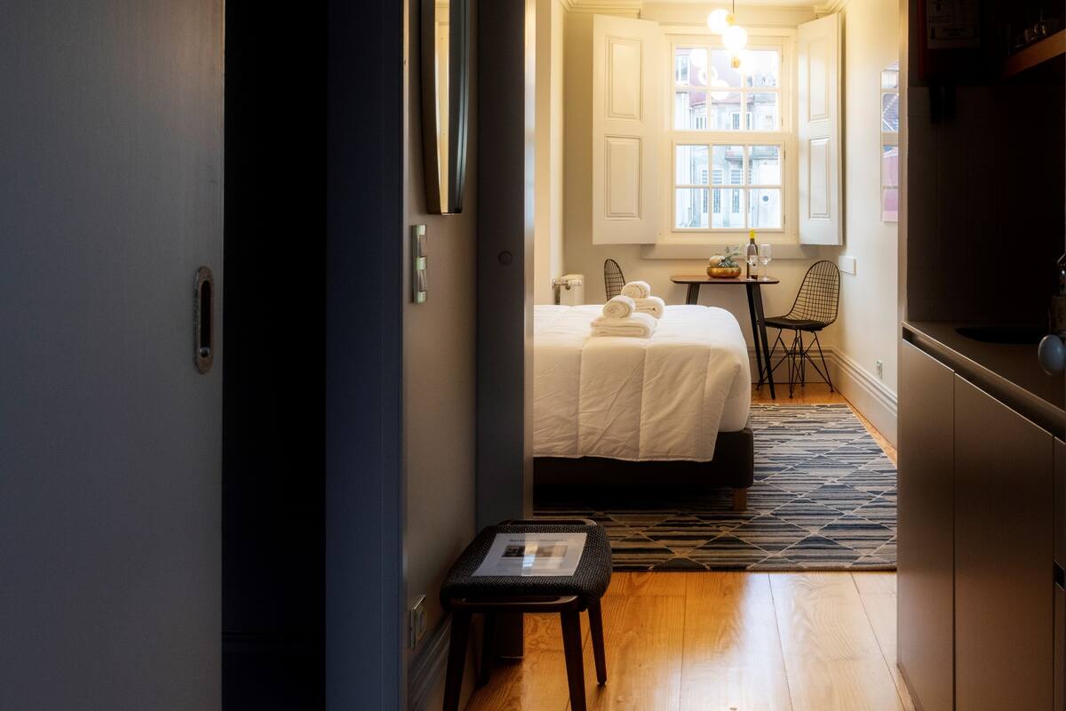 A hotel room with a bed, a small table, a chair, and a window with curtains.