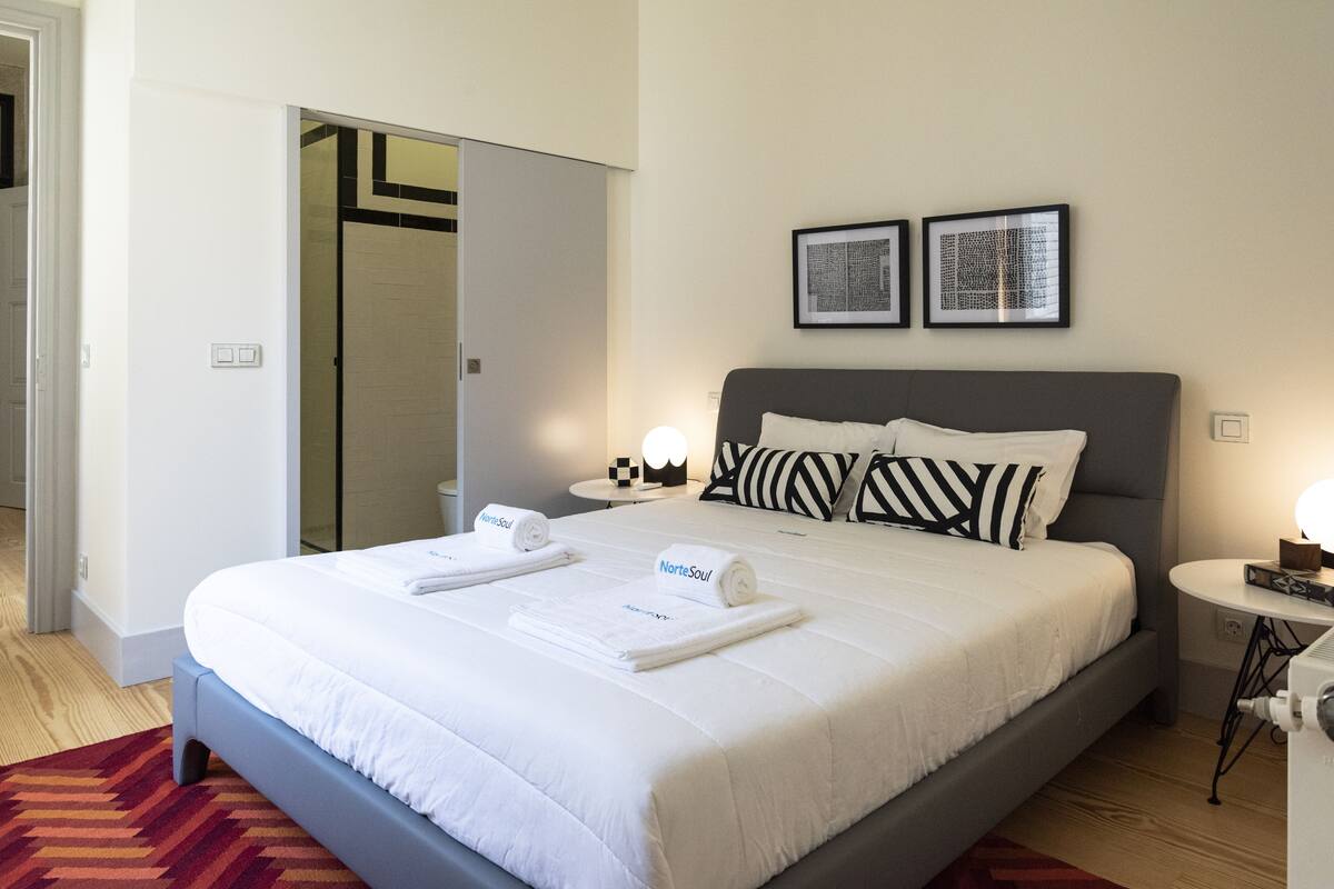 A neatly made bed with white bedding and rolled towels, a grey headboard, and two framed pictures on the wall.