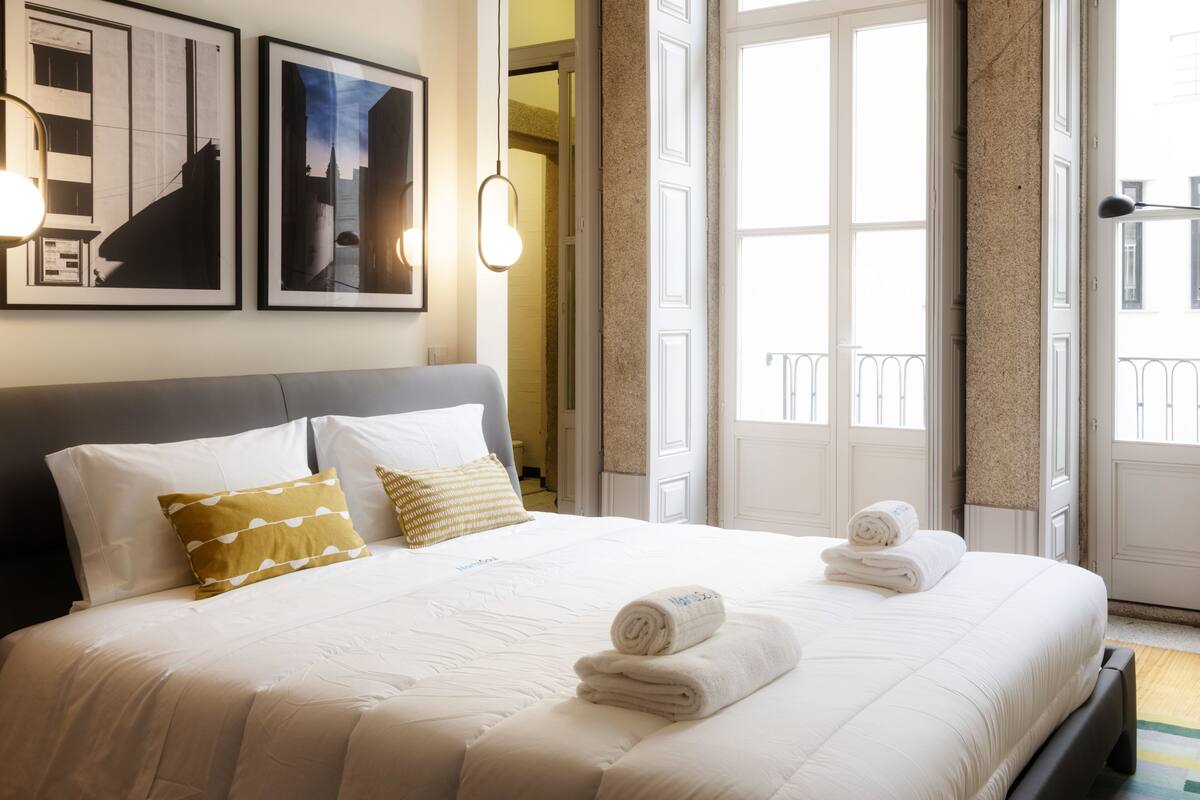 A neatly made bed with white linens and two pillows, a framed picture above the bed, and a window with curtains.
