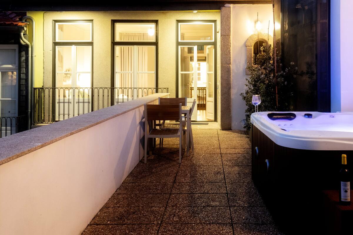 A balcony with a hot tub, a table, and chairs, and a building with illuminated windows and a dome structure in the background.