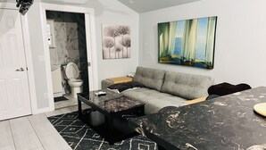 Una sala de estar moderna con un sofá gris, una mesa de centro negra y una mesa de comedor con diseño de mármol. Un cuadro de un paisaje cuelga en la pared, y se ve un baño al fondo.