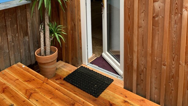 Pared exterior de madera con planta en maceta y felpudo negro en la entrada.