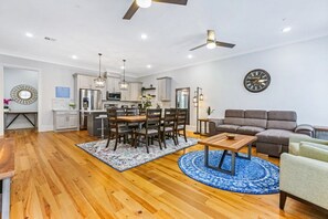 A modern kitchen with white cabinets, a dining area with a wooden table and chairs, a living room with a sofa and a coffee table, and a clock on the wall.