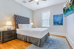 A bedroom with a tufted headboard bed, two bedside tables with lamps, a large window with blinds, and a wall-mounted TV displaying 'Hosteva'.