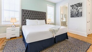 Un dormitorio con cabecera capitonada, una cama grande con ropa de cama blanca, una alfombra estampada y una mesita de noche con lámpara.