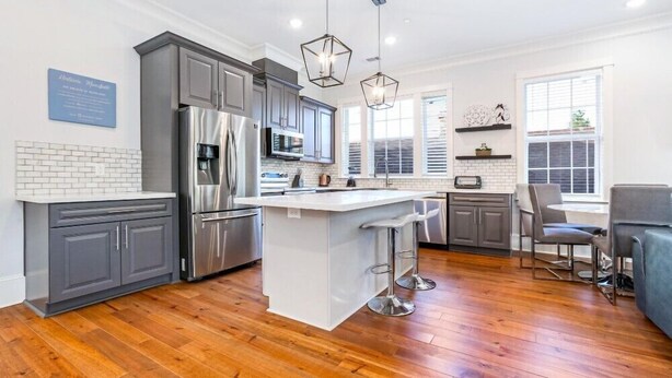 A modern kitchen with grey cabinets, stainless steel appliances, a central island with bar stools, and pendant lighting. - 15% OFF Monthly | 10% OFF Weekly- Chic Urban Retreat w Pool Steps to St. Charles Ave and (New Orleans)