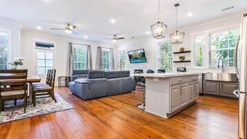 A spacious living area with a grey sectional sofa, wooden dining table, and chairs. There is a kitchen with a white island, stainless steel appliances, and a large window.