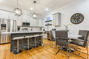 A modern kitchen with white cabinets, stainless steel appliances, a bar area with stools, and a dining table with chairs. - 5BR w Pool | Great for Families | Near Everything (New Orleans)