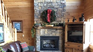 L’intérieur d’un chalet accueillant, avec une cheminée en pierre, des murs en bois, un canapé en cuir et un téléviseur posé sur un meuble en bois.