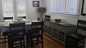A dining area with a table set for four, surrounded by chairs, a cabinet with drawers, and a window with blinds.