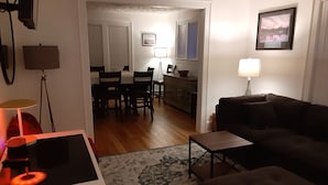 A living room with a dark brown sofa, a small wooden coffee table, and a patterned rug. There is a dining area with a table and chairs in the background.