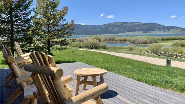 Una terraza de madera con sillas Adirondack y una pequeña mesa redonda, con vistas a un paisaje pintoresco con un lago y montañas.