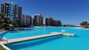 Piscina com um deck, rodeada por edifícios altos, palmeiras e um céu claro.