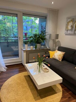 A modern living room with a sofa, a coffee table with a vase of flowers, and a view of greenery outside.