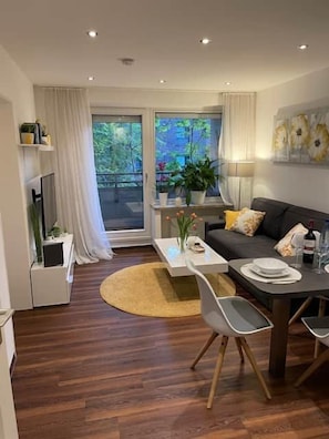 Un salon moderne avec un canapé, une table basse et une table à manger. Des plantes sont posées sur le rebord de la fenêtre et un téléviseur est installé sur un meuble.