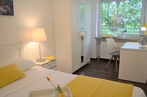 A hotel room with a bed, bedside table, lamp, desk with chair, and a window with a view of greenery.